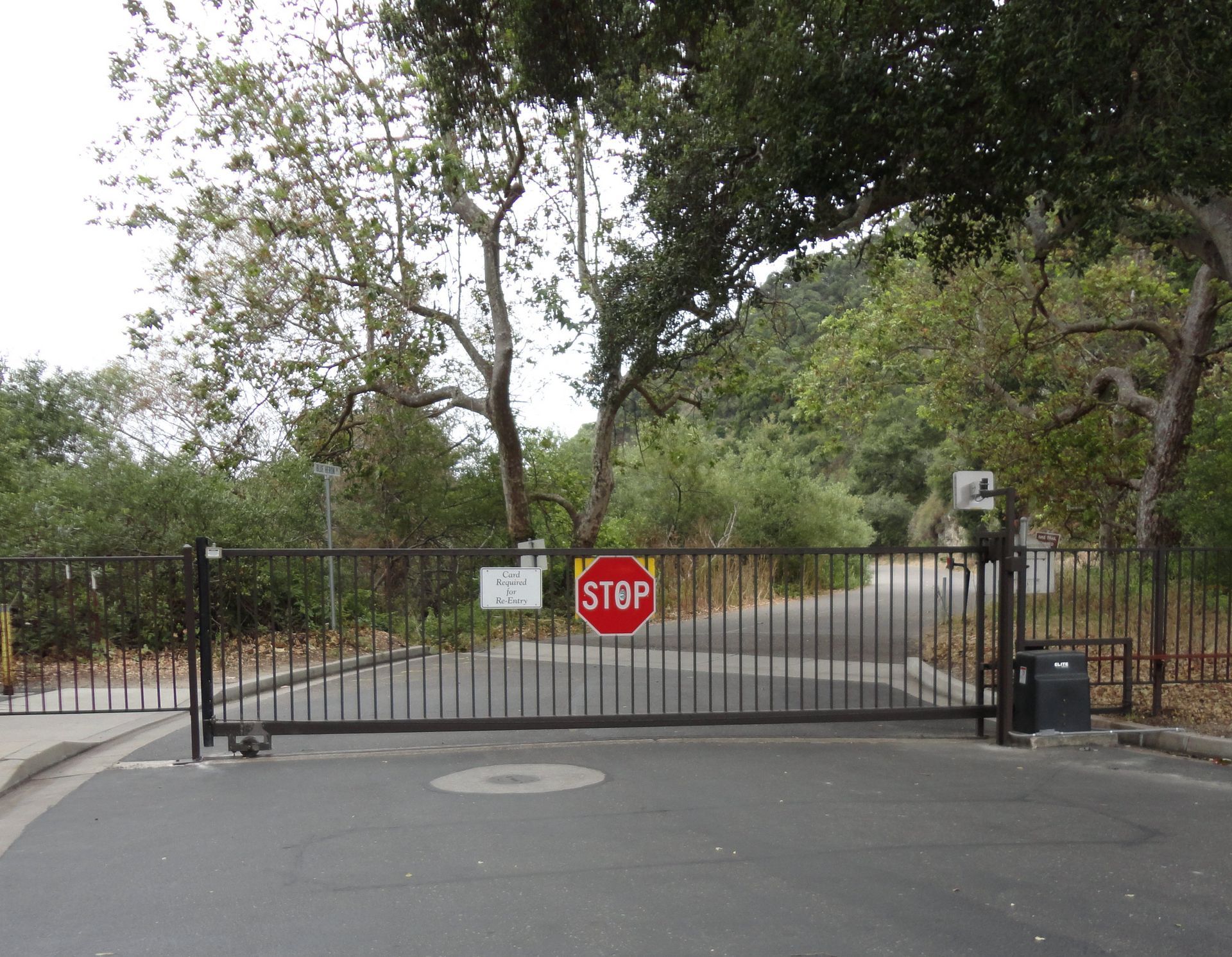 Black metal gate with STOP sign blocks a tree-lined road. A security camera and a trash can are present.
