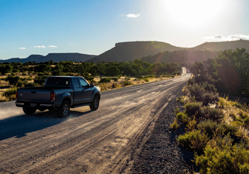 Blue pickup truck driving down a dusty desert road at sunset with mountains and open landscape.