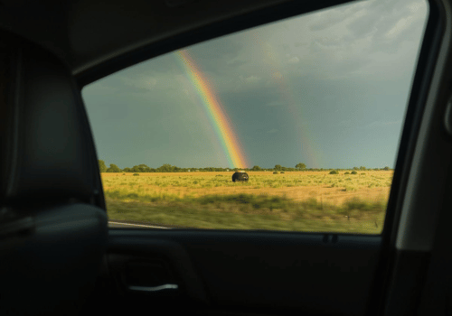 View through a car window of an open field with a bright rainbow in the distance.