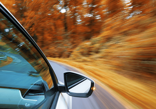 Car speeding along a curving road through autumn forest, seen from the side mirror view.