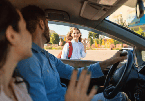 Driver smiling as a student boards a car, with two passengers inside and a suburban street outside