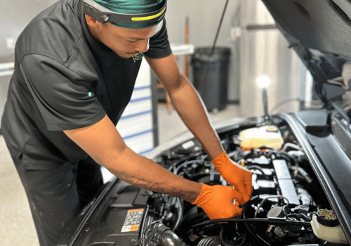 Mechanic in black uniform and orange gloves working under a car hood in a garage