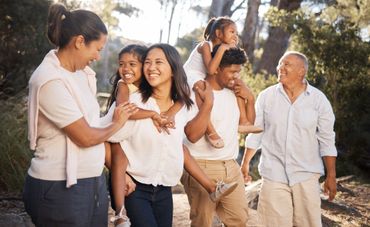 A smiling family walking together outdoors in warm sunlight, with two children carried by adults.