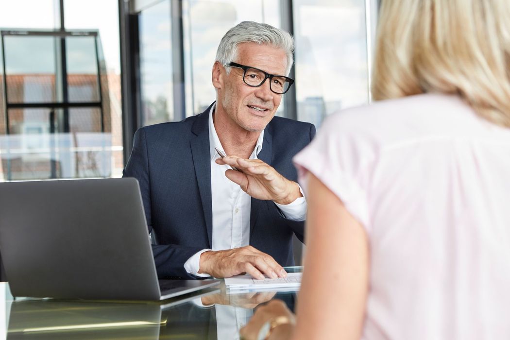 Business meeting with a smiling man gesturing across a glass desk with a laptop in a bright office