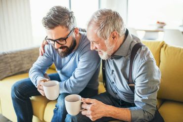 Two people sit on a couch sharing coffee and looking at something together in a bright living room.