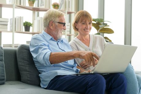 Two people sitting on a couch, smiling and using a laptop in a bright living room.