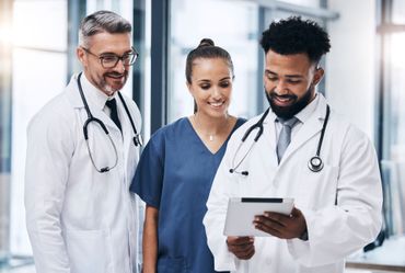 Three doctors in white coats reviewing a tablet together in a bright hospital hallway