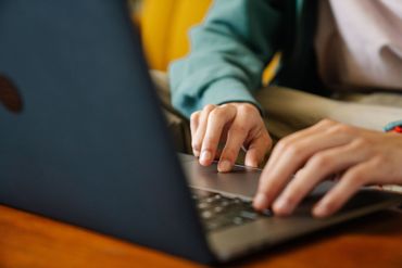 Hands typing on a laptop at a wooden table, with a blurred blue and yellow background.
