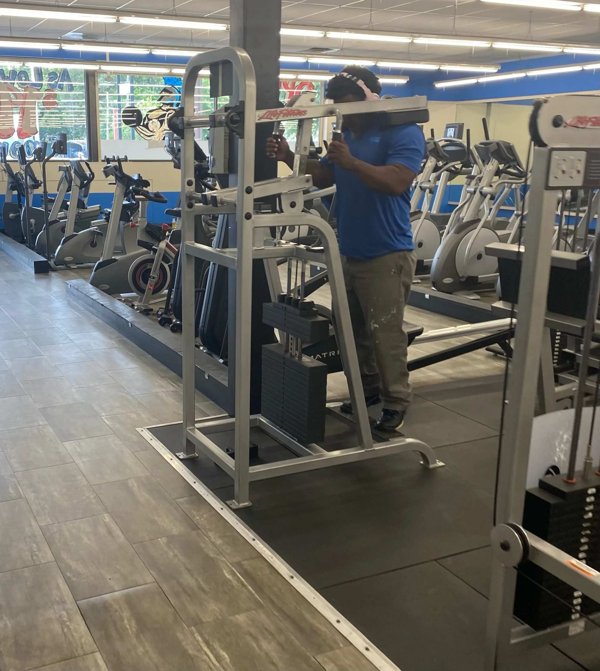 A person using a standing squat machine in a gym. Grey machine, blue shirt, gray pants. Other equipment visible.