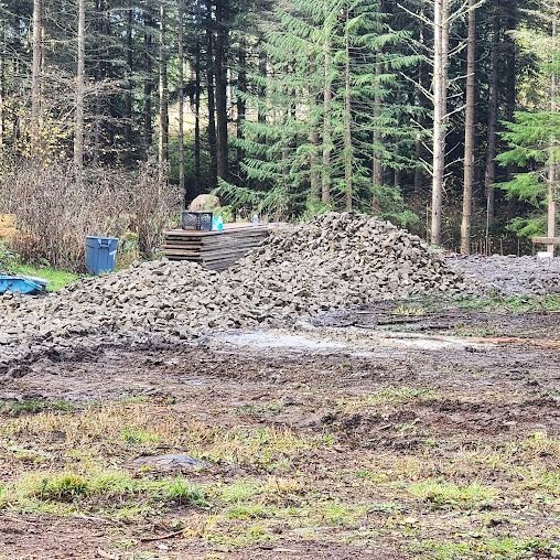 Pile of gravel and dirt in a forest clearing with pine trees and a blue bin nearby