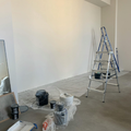 Empty room with white walls, drop cloths, paint buckets, and two ladders during painting or renovation.
