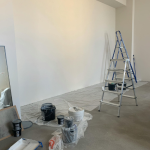 Empty room with white walls, drop cloths, paint buckets, and two ladders during painting or renovation.