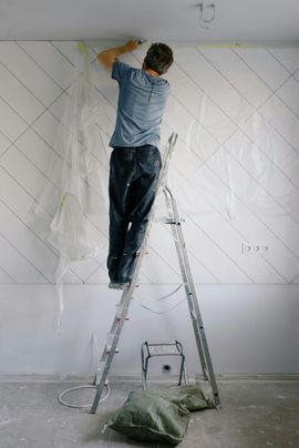 Person standing on a ladder painting a white wall in a room under renovation.