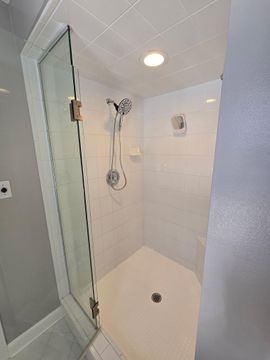 Modern white tiled shower stall with glass door, built-in bench, handheld showerhead, and floor drain