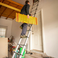 Person on a ladder carrying yellow boards while working near a white door in a garage.