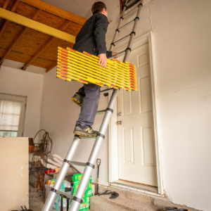 Person on a ladder carrying yellow boards while working near a white door in a garage.