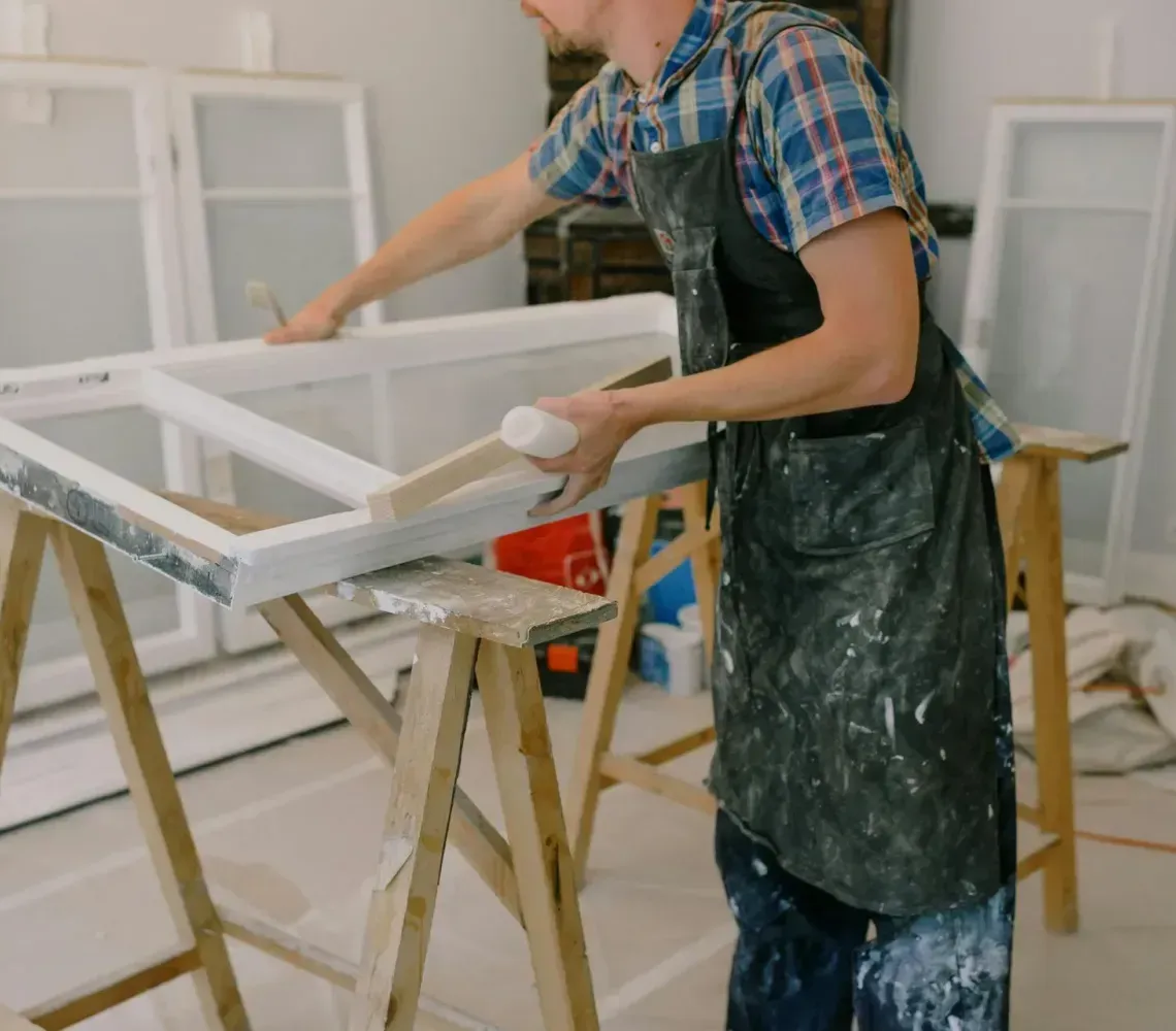 Person painting a white window frame on sawhorses in a room under renovation