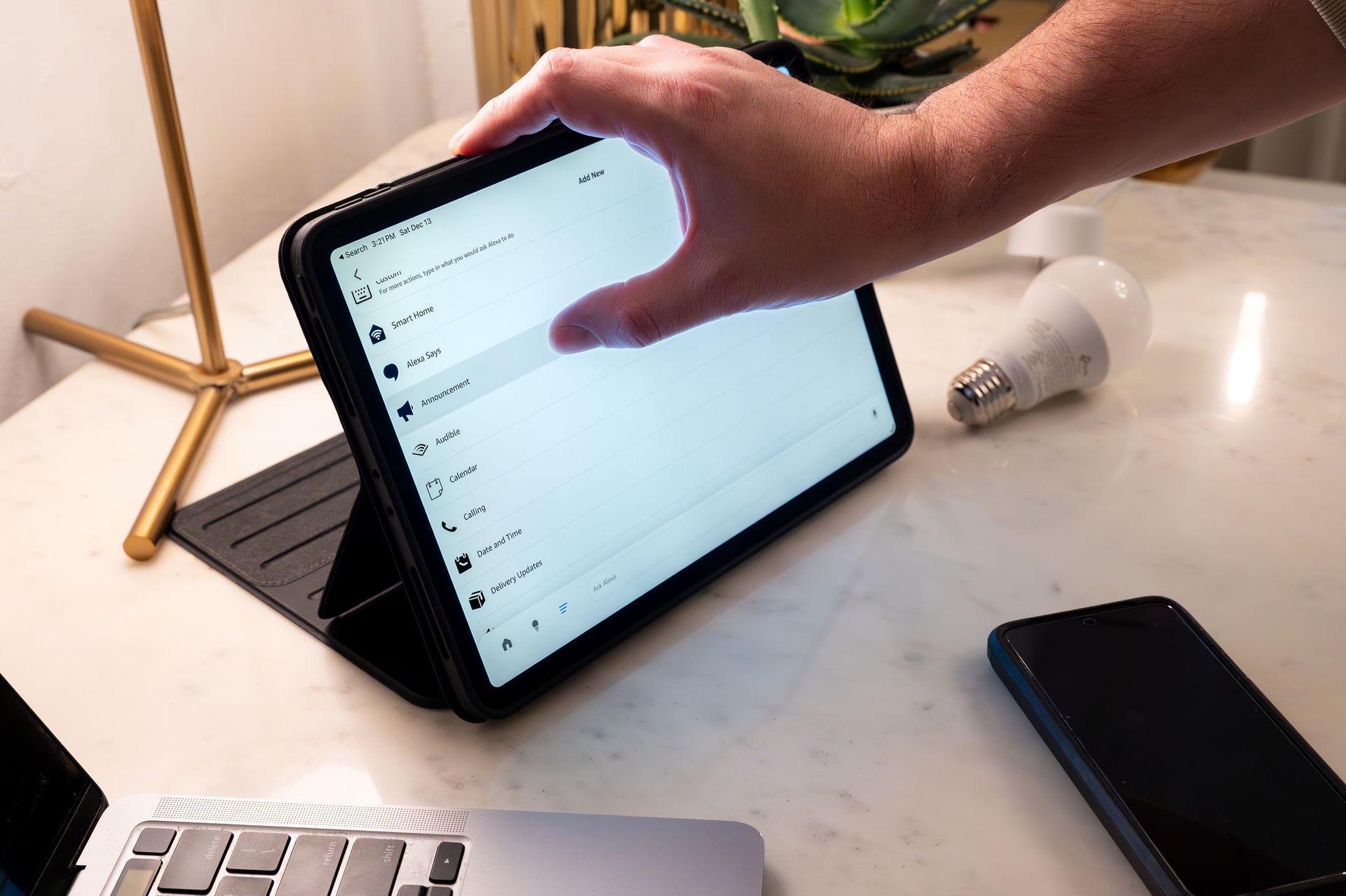 A hand touching a tablet screen on a stand.  A lightbulb and phone are also on the table.