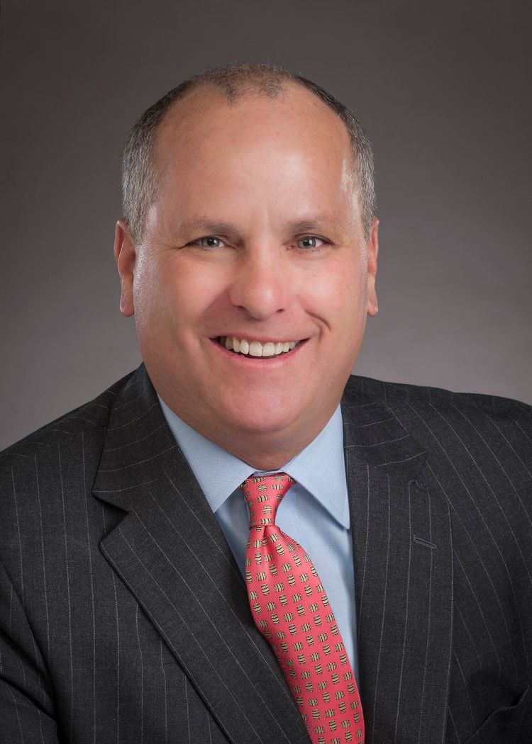 Headshot of a smiling man in a dark suit and red tie against a neutral gray background