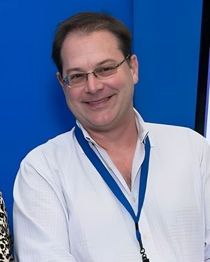 Smiling man in a white shirt with blue lanyard standing against a blue background