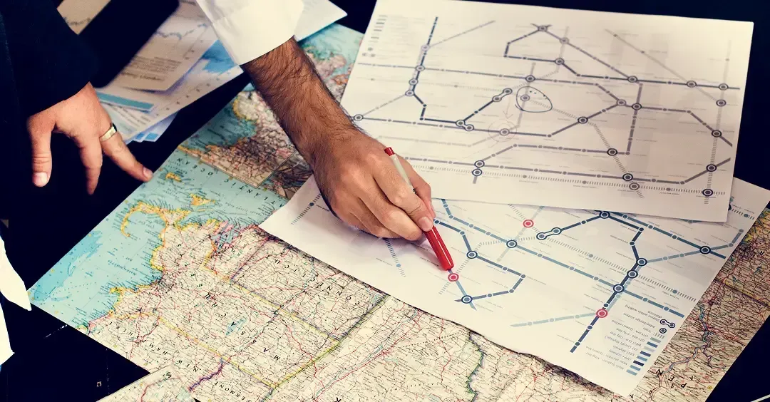 Hands pointing at paper maps and route sketches on a table, marking locations with a red pen.