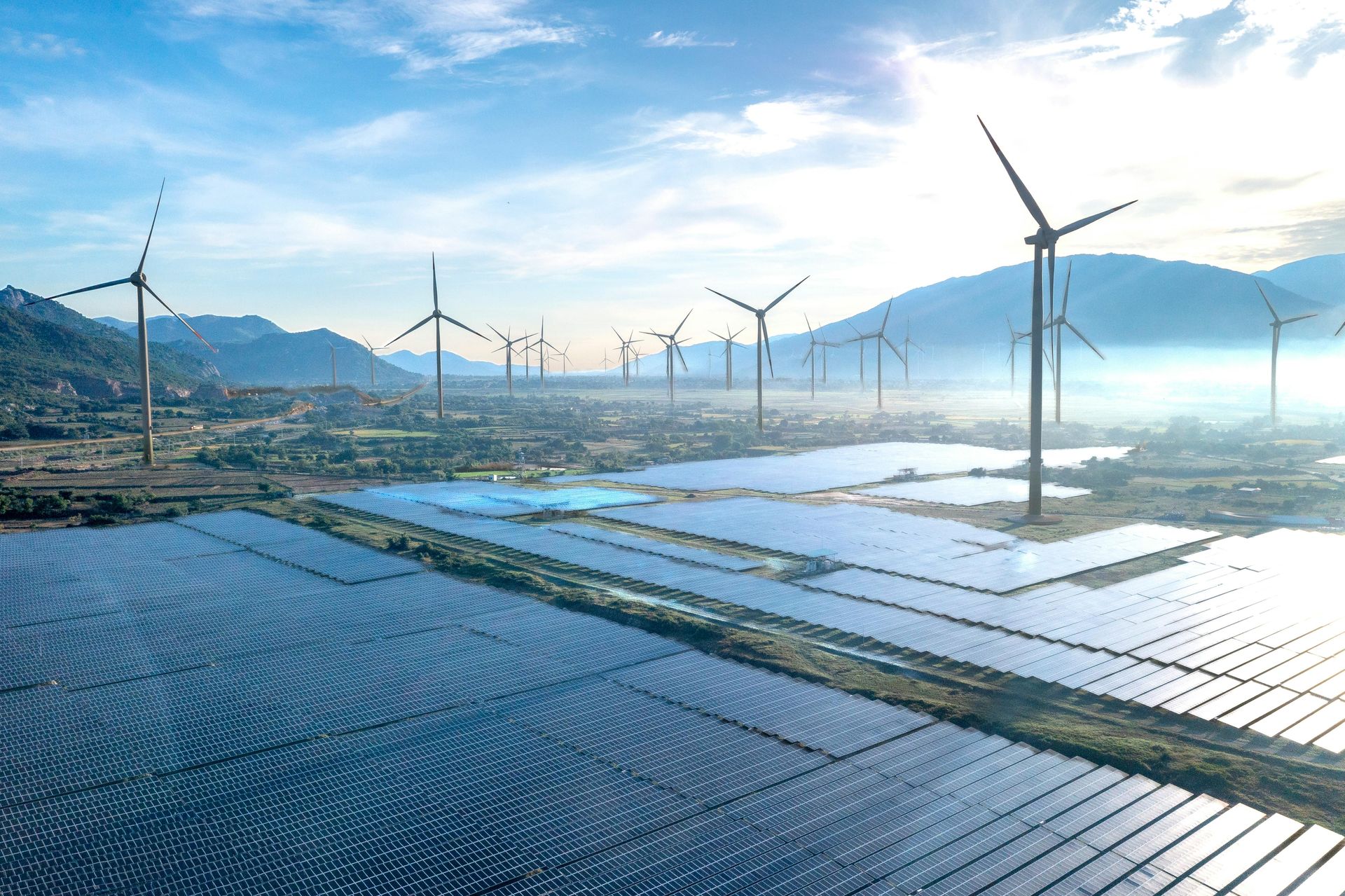 Wind turbines overlooking a solar farm with water channels and mountains in the distance