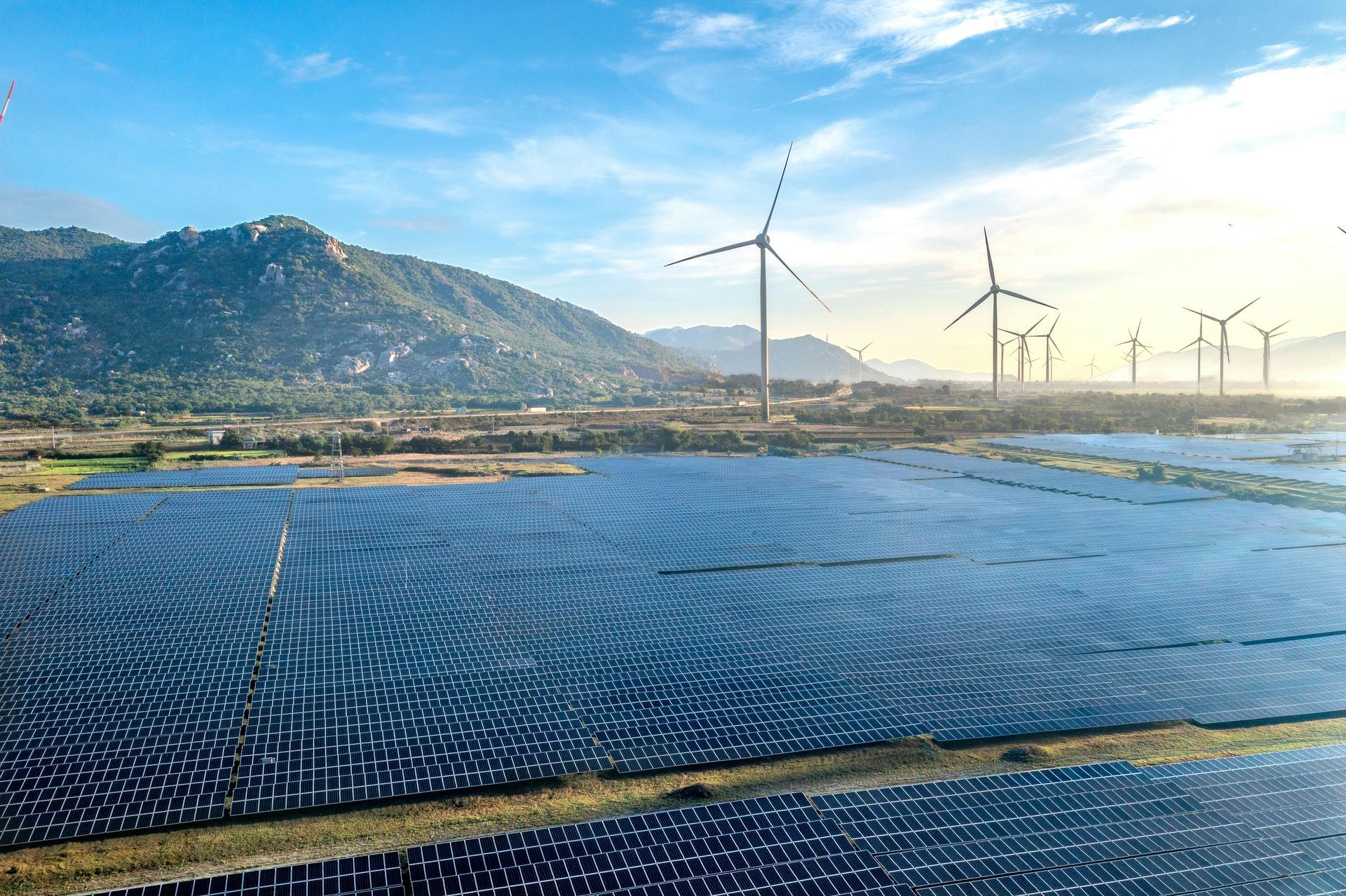 Solar panels in the foreground with wind turbines, mountains, and a blue sky in the background.