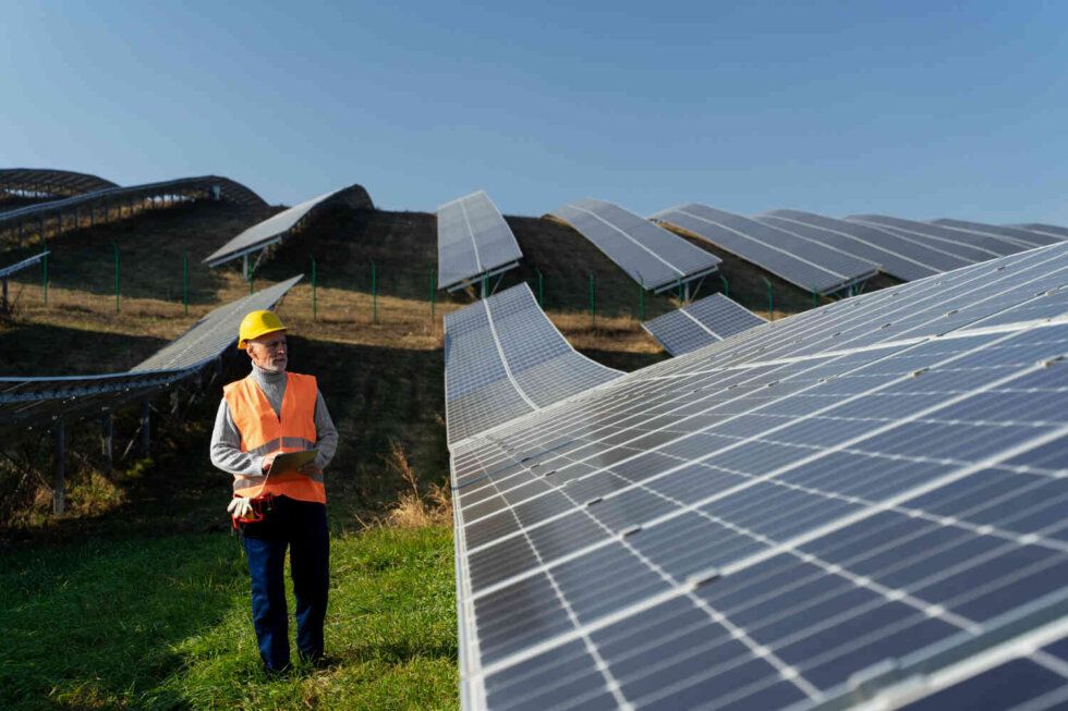 Worker in orange safety vest and hard hat inspecting rows of solar panels on a hillside