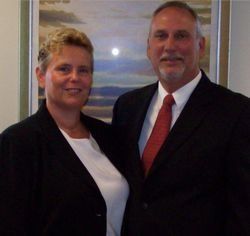 A person with short blonde hair and a person with a goatee in a suit and red tie pose together in front of a landscape art.