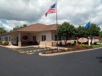 A single-story building with a brown roof and American flag, featuring a paved parking lot and a sign in the front lawn.