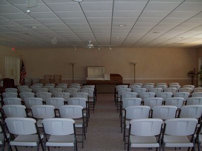 A viewing room featuring rows of gray chairs facing an open casket on a pedestal, with an American flag to the left.