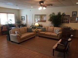 A waiting area with two beige sofas, an armchair, a table lamp, and potted plants on a tiled floor.