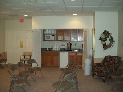 A room with a small kitchenette, coffee station, and beige folding chairs arranged for a meeting or gathering.