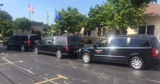 A row of three black funeral vehicles parked in a paved lot in front of a building with two flags flying.