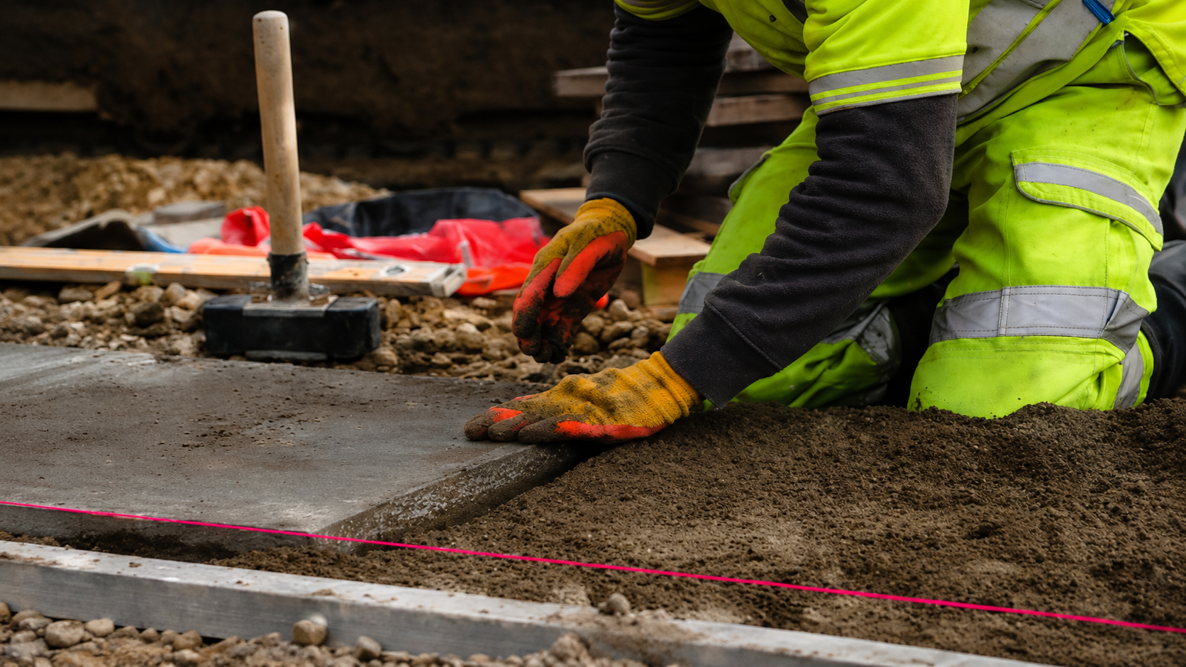 Construction workers spreading concrete on a sidewalk with trowels, wearing neon safety gear.