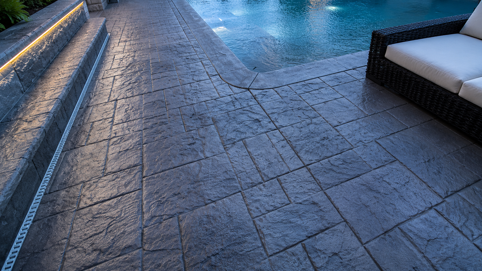 Stone pool deck beside a blue-lit swimming pool, with a cushioned lounge chair on the right.