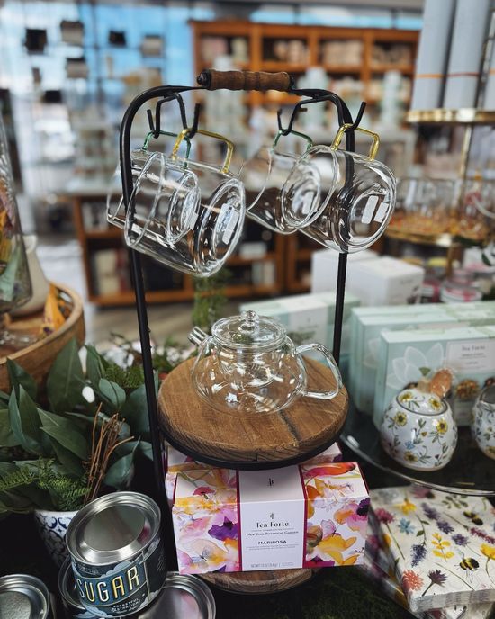 Glass teapot and cups on a black metal stand in a shop, with tea boxes and floral decorations.