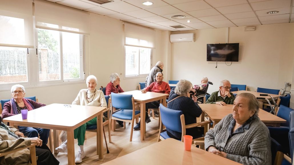 Persone sedute ai tavoli in una luminosa sala comune, con un televisore a parete.