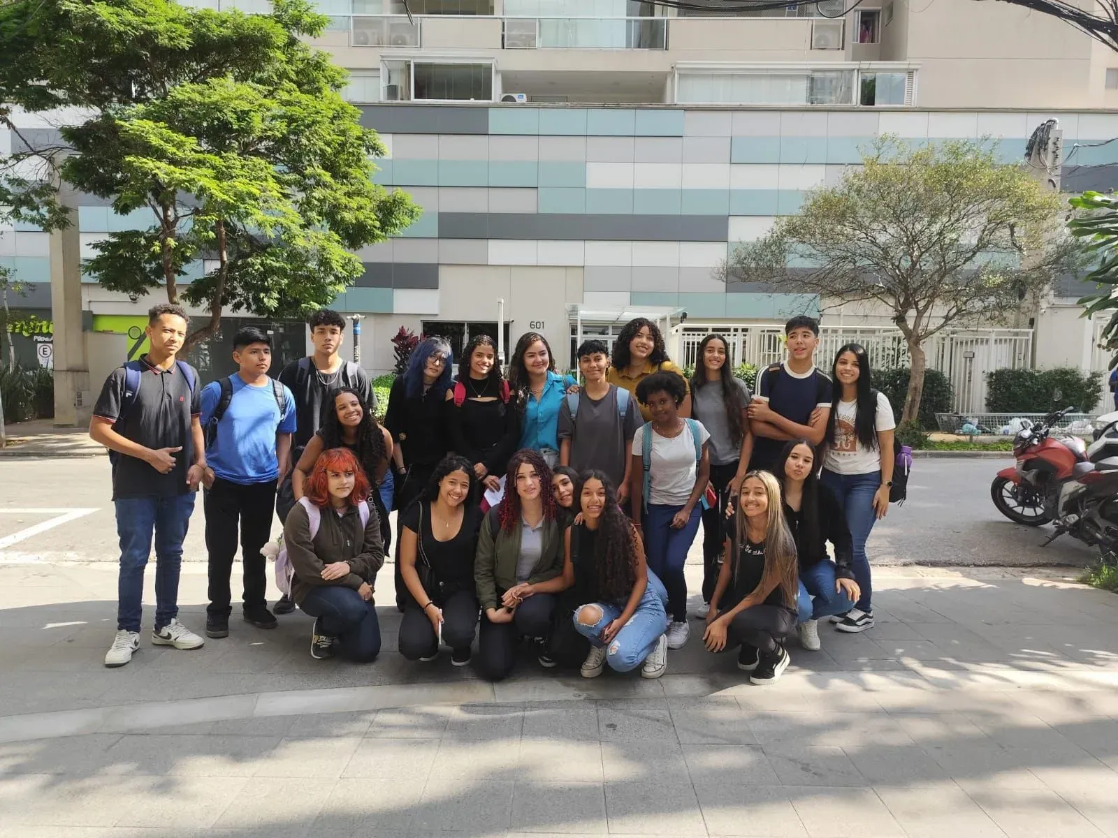 Um grande grupo posando ao ar livre em frente a um prédio moderno, com árvores e motocicletas estacionadas nas proximidades.