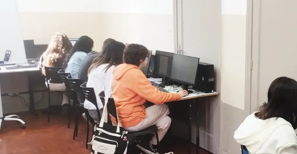 Pessoas sentadas em computadores de mesa em um laboratório de informática bem iluminado, trabalhando em silêncio.
