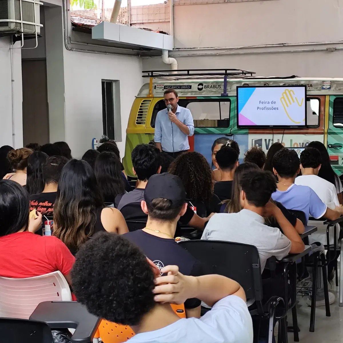 Público sentado para uma palestra em um pátio externo, palestrante na frente