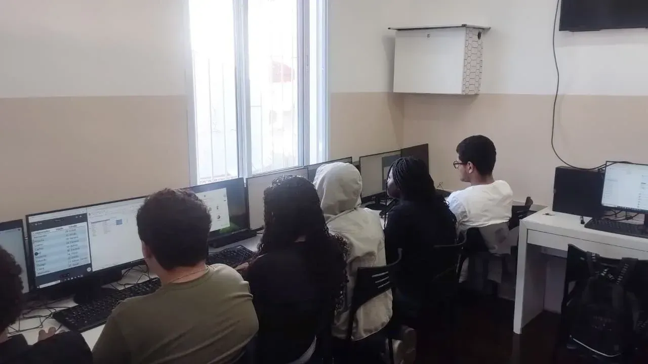 Pessoas sentadas em frente a computadores em uma sala de aula ou laboratório de informática