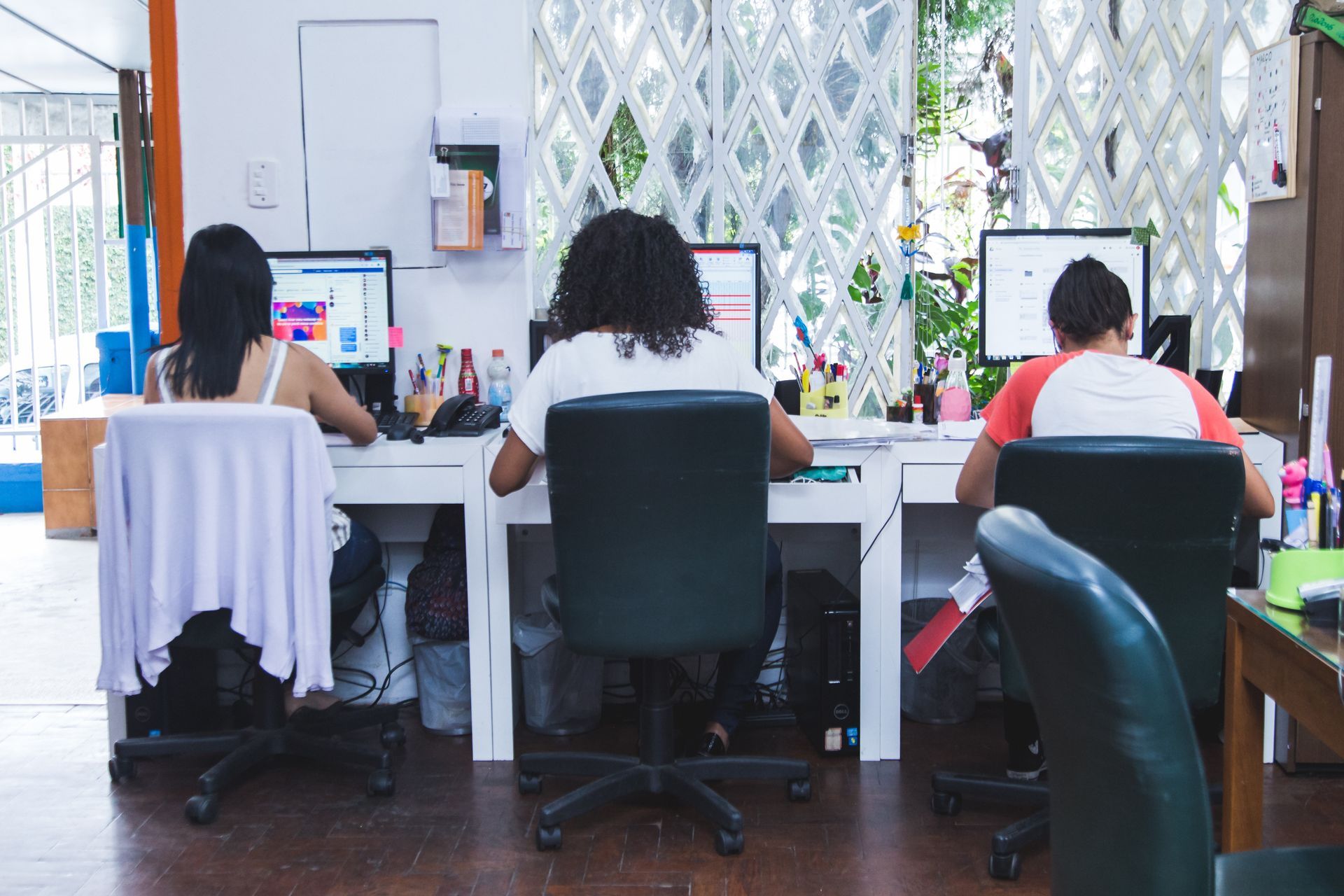 Pessoas trabalhando em computadores de mesa em um escritório com grandes janelas e decoração de parede estampada.
