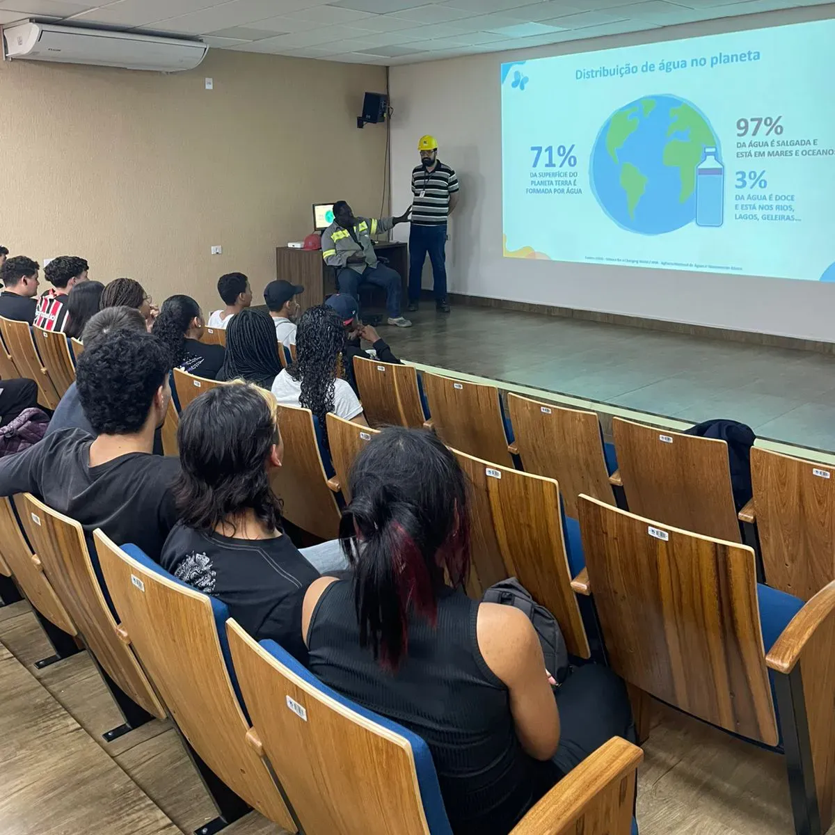 Público sentado em um auditório assistindo a uma apresentação com slides projetados.