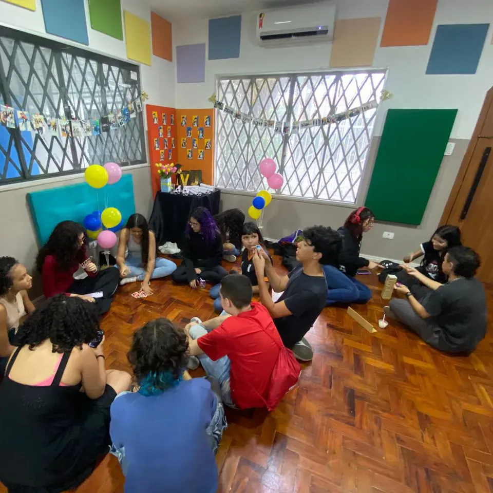 Pessoas sentadas em círculo sobre um piso de madeira, conversando em uma sala colorida com balões e janelas quadriculadas.