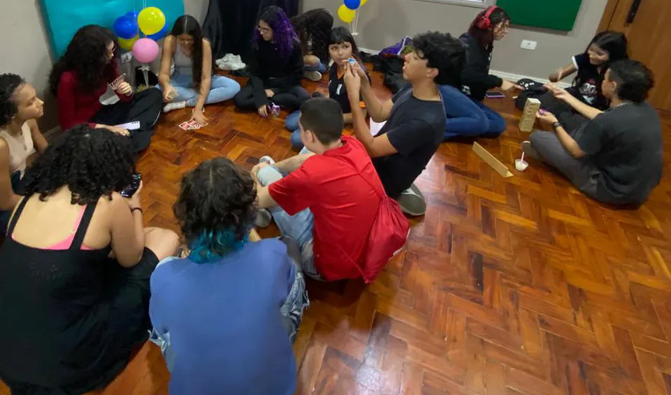 Pessoas sentadas em círculo no chão de madeira, reunidas em torno de balões
