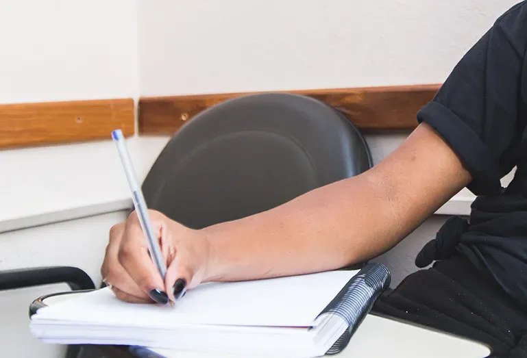 Pessoa escrevendo em um caderno sentada em uma cadeira em um escritório.