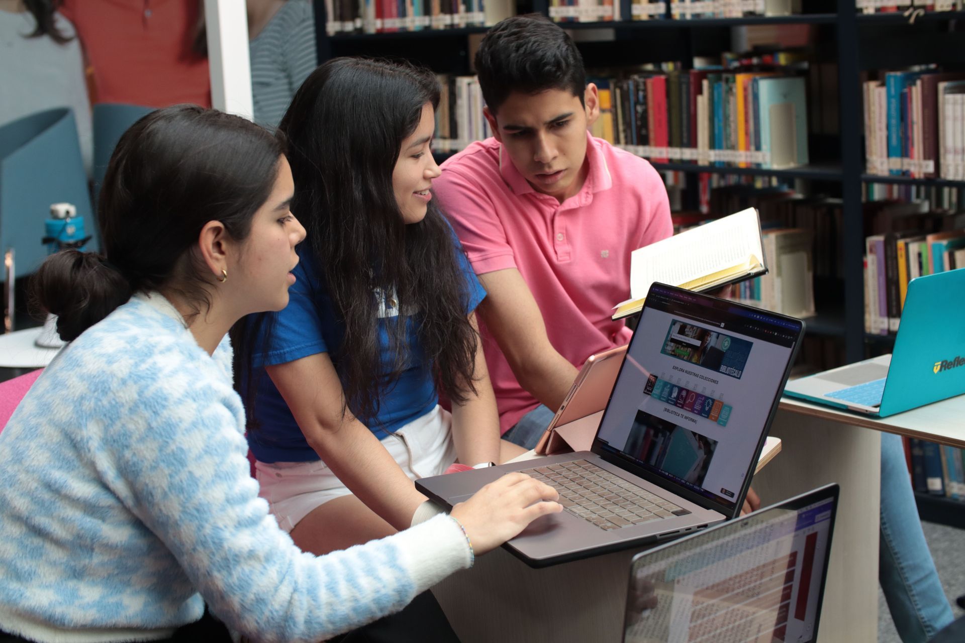 Três estudantes colaborando em torno de um laptop em uma biblioteca, com um deles apontando para a tela.