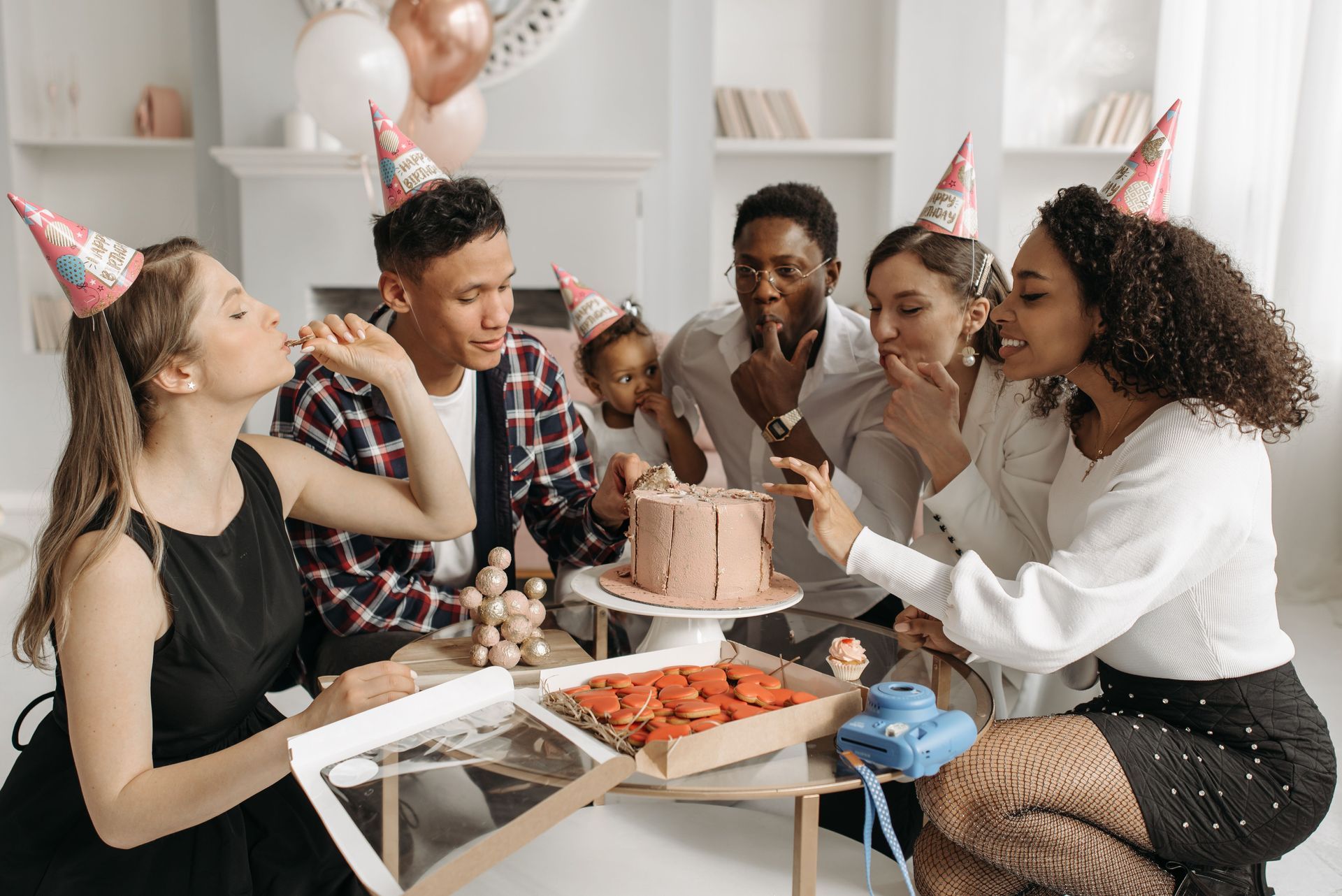 Amigos com chapéus de festa se reúnem em volta de um bolo de aniversário em uma mesa, sorrindo e conversando dentro de casa.