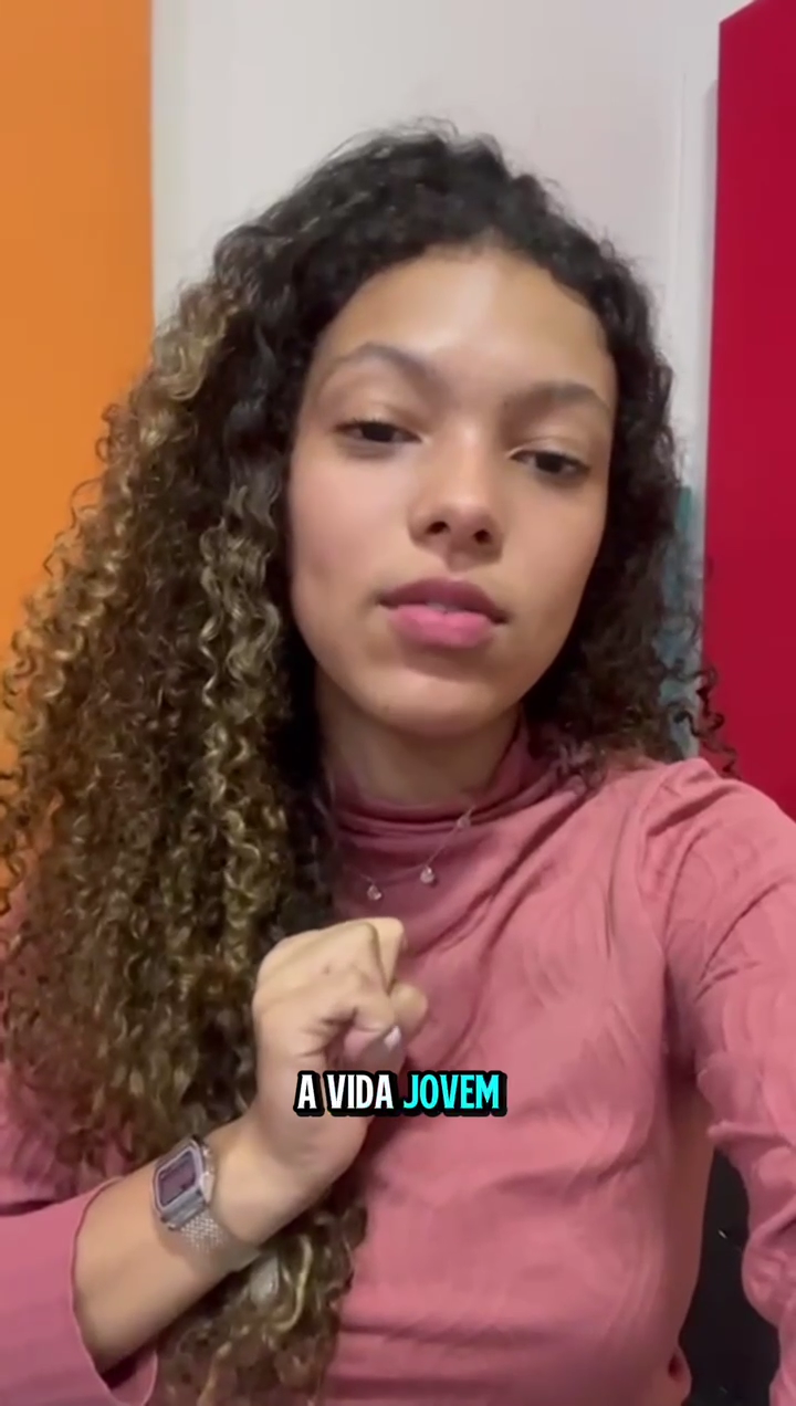 Mulher de blusa rosa tirando uma selfie em ambiente interno, com cabelos cacheados e expressão neutra.